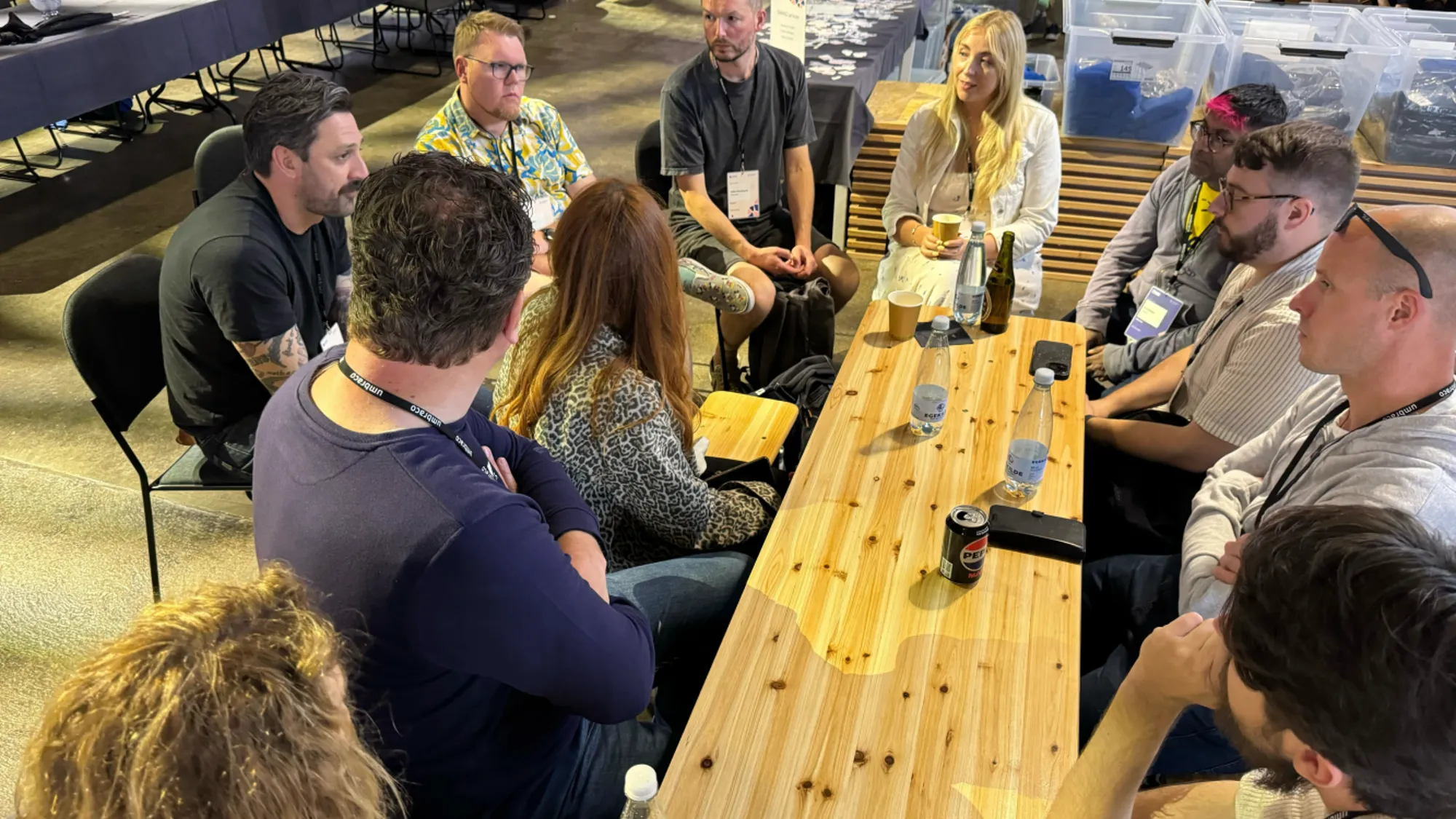 A wooden picnic bench style table surrounded by people. Some are facing the camera and some have their backs to the camera.