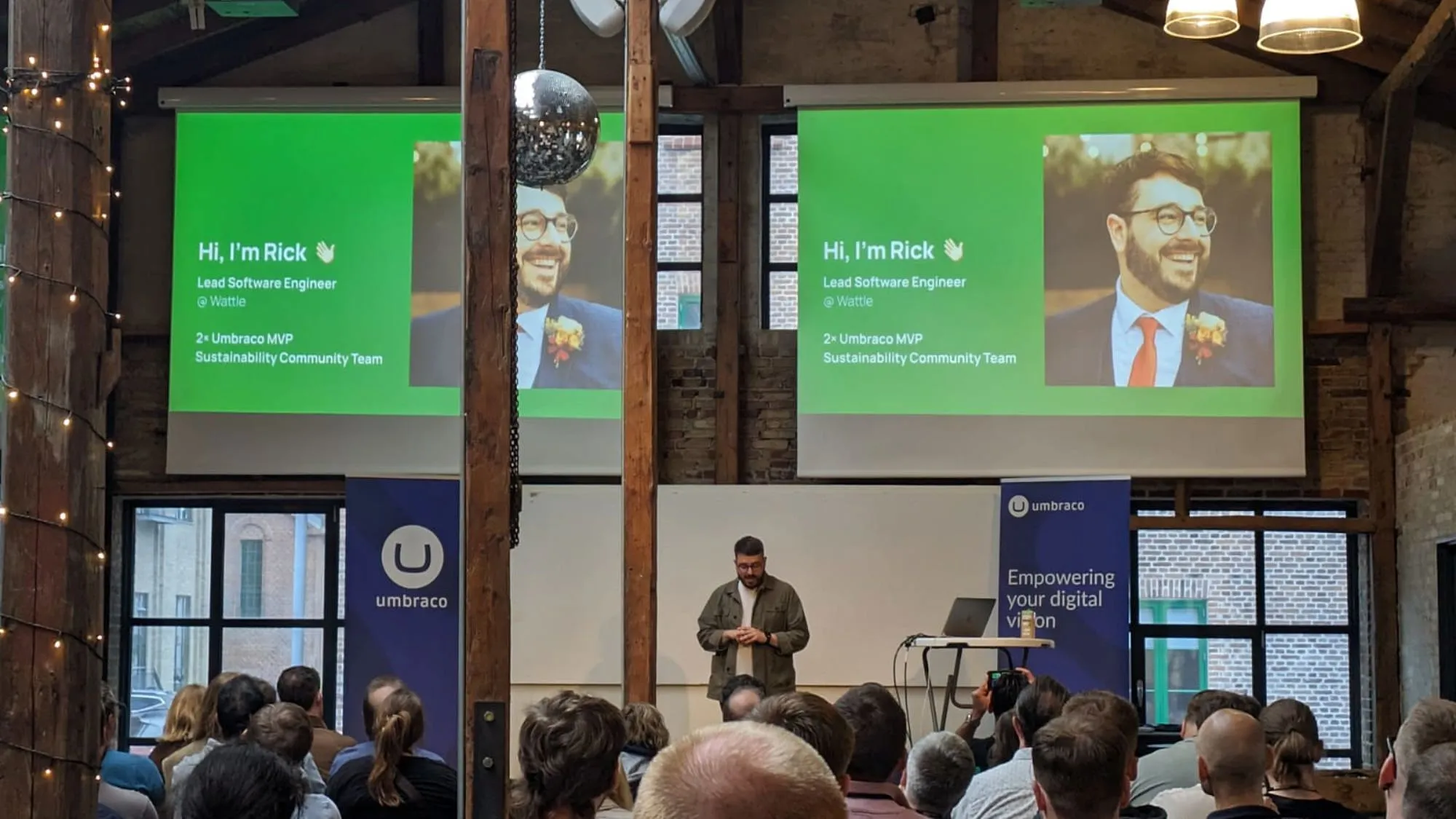 A full conference room with two large projector screens on the back wall, featuring a bright green slide, Rick's picture and text introducing Rick. He is stood on the stage at the bottom.