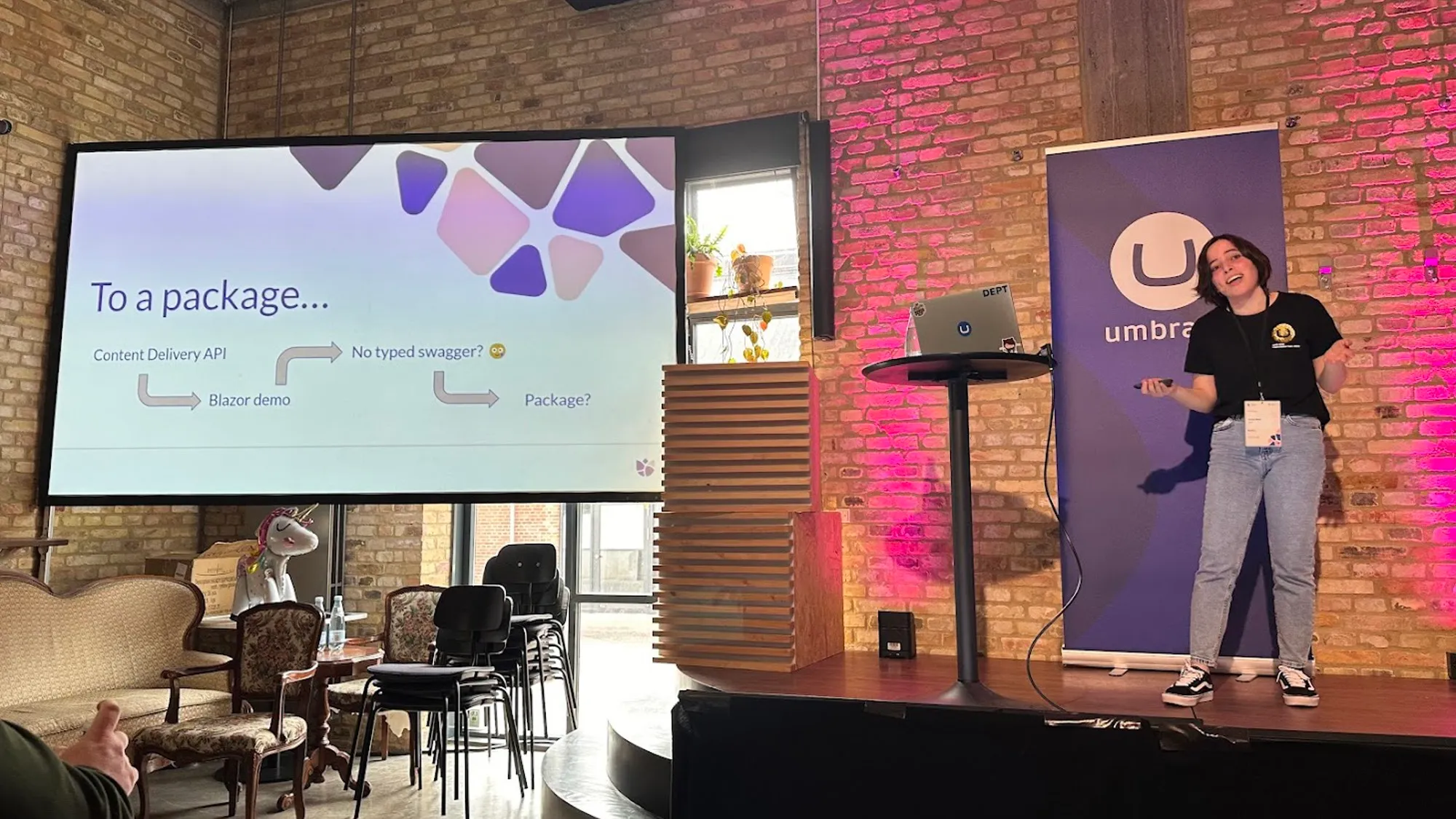 Laura Neto on stage giving her talk. Behind her is a pull-up blue Umbraco banner and a brick wall illuminated by pink lights. To the left is a projector screen showing her slides.