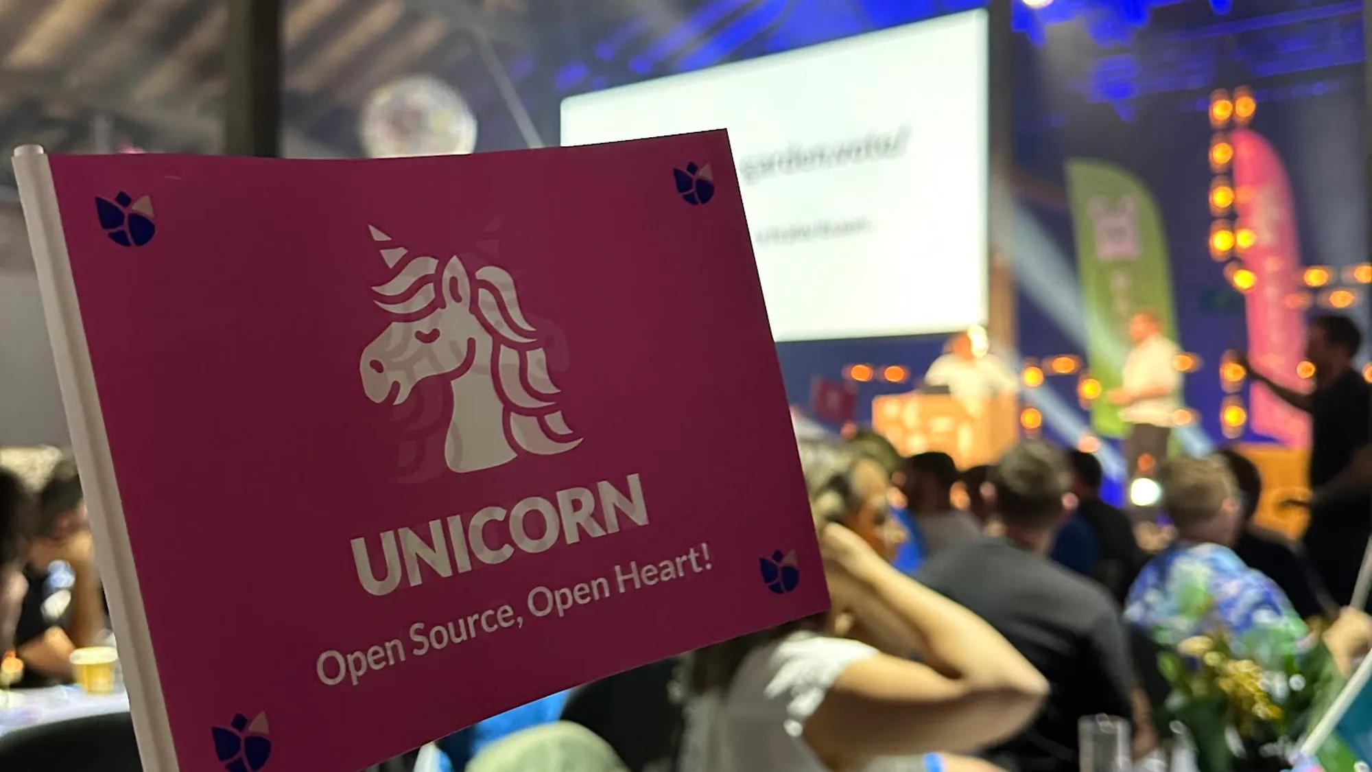 A pink flag with an illustration of a unicorn, reading "Unicorn. Open source, open heart!". In the background is the Codegarden conference out of focus